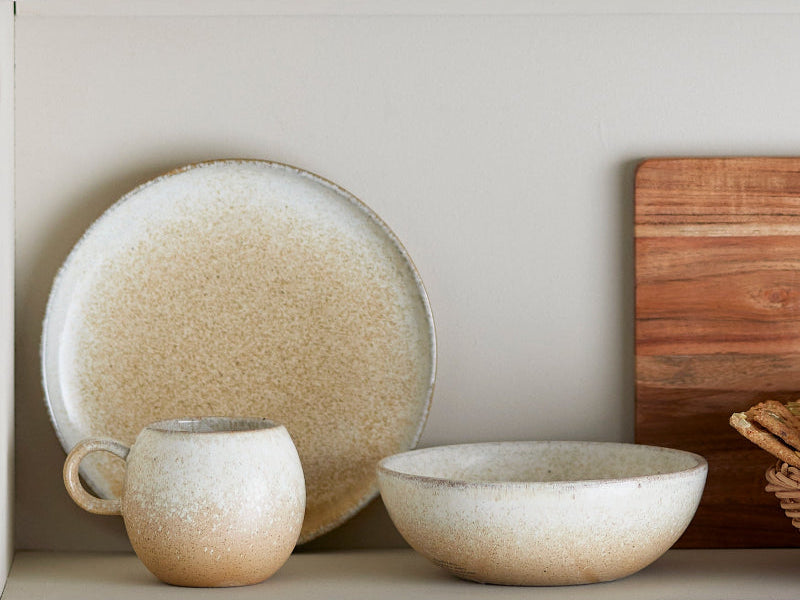 Set of ceramic dinnerware including plates, cups, and bowls on a shelf with a wooden cutting board.