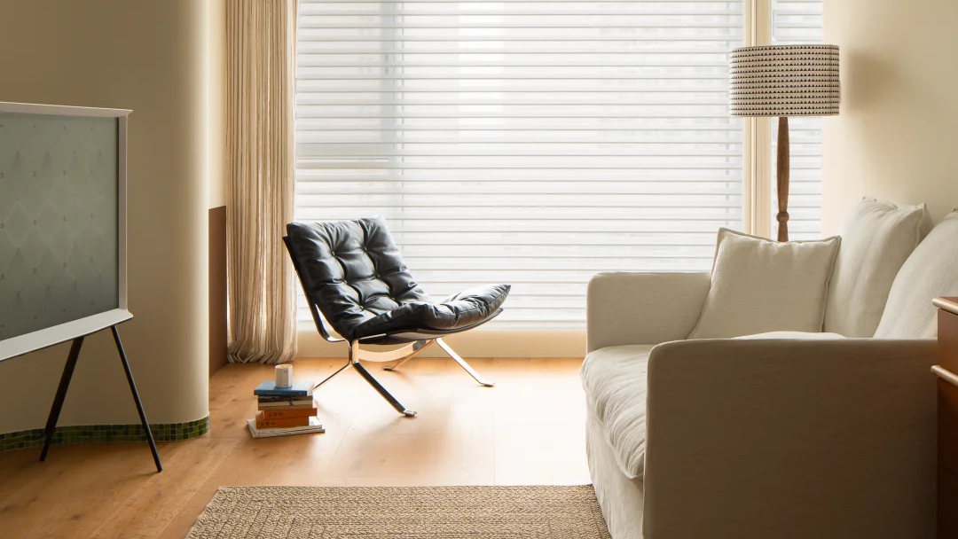 A modern living room corner featuring a cream-colored fabric sofa, a black leather lounge chair, a woven area rug, and a large window with white blinds and beige curtains.