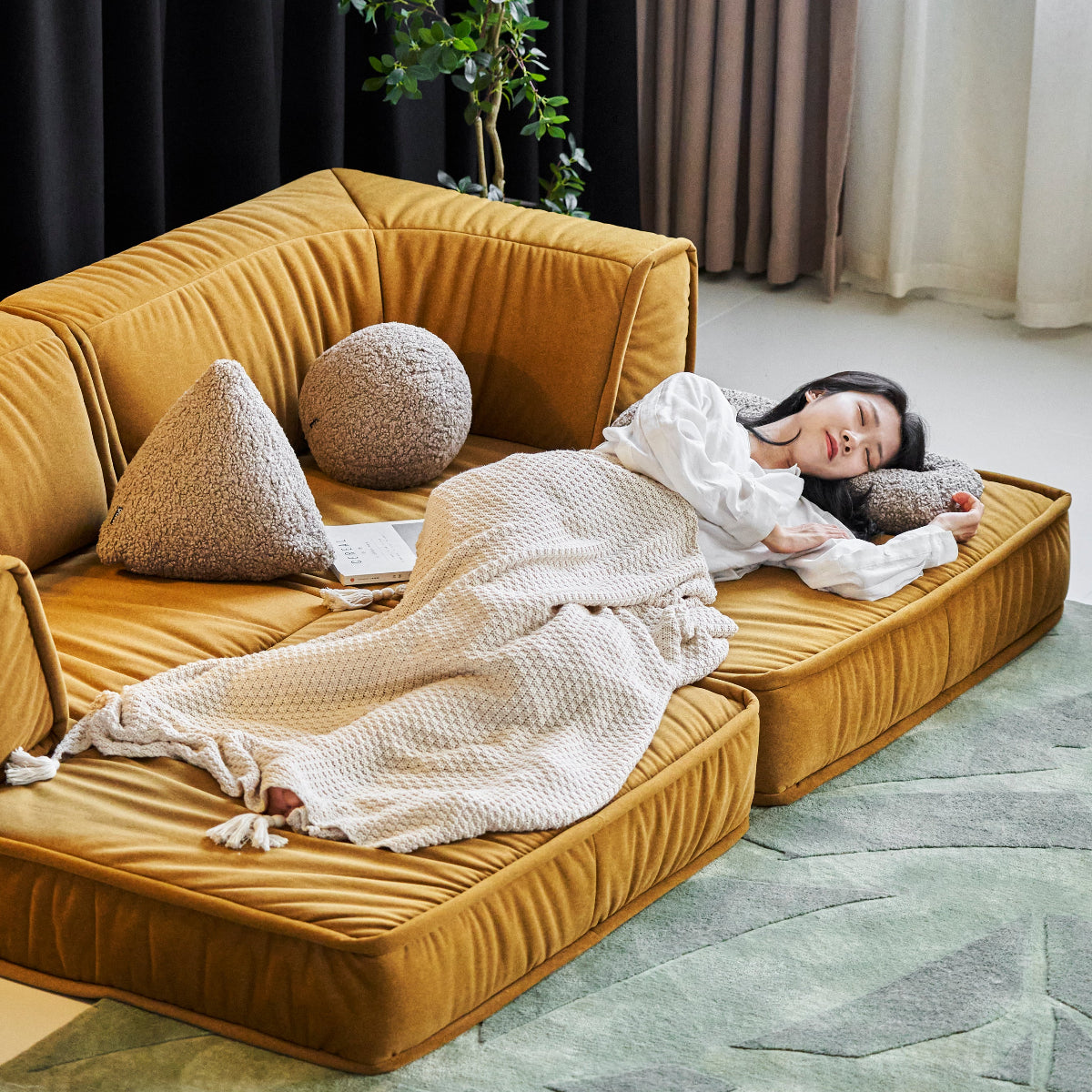 Person lying on a mustard yellow sofa with decorative pillows and a blanket in a modern room.