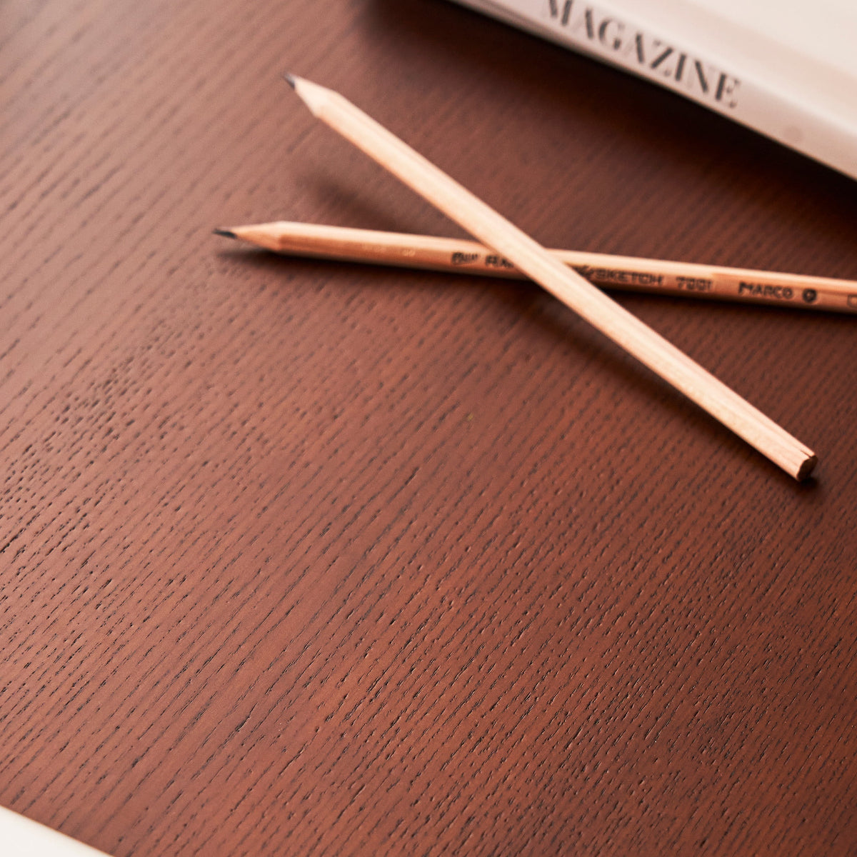 Two pencils on a wooden surface with a magazine in the background