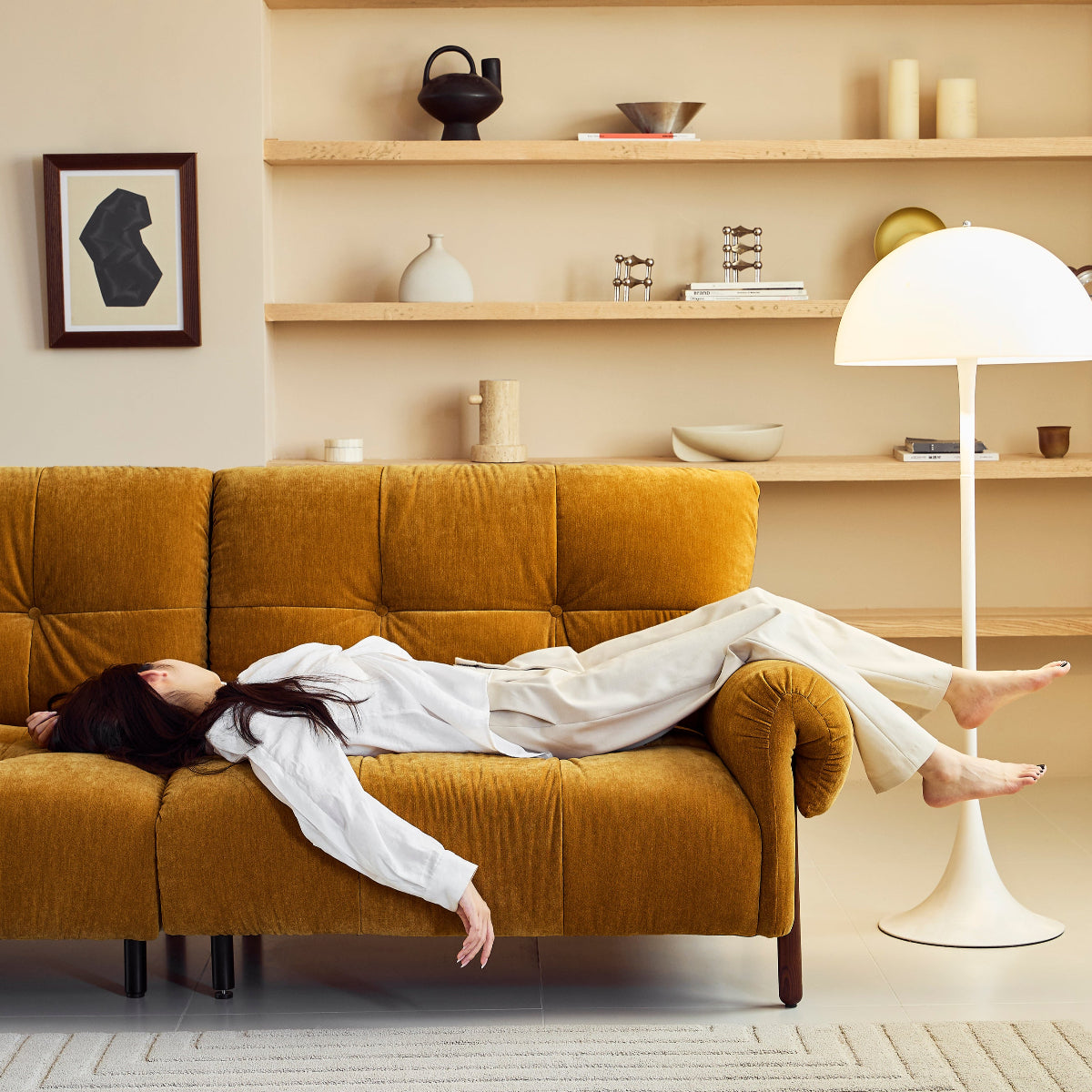 Person lying on a mustard yellow sofa in a modern living room with wooden shelves and a floor lamp.