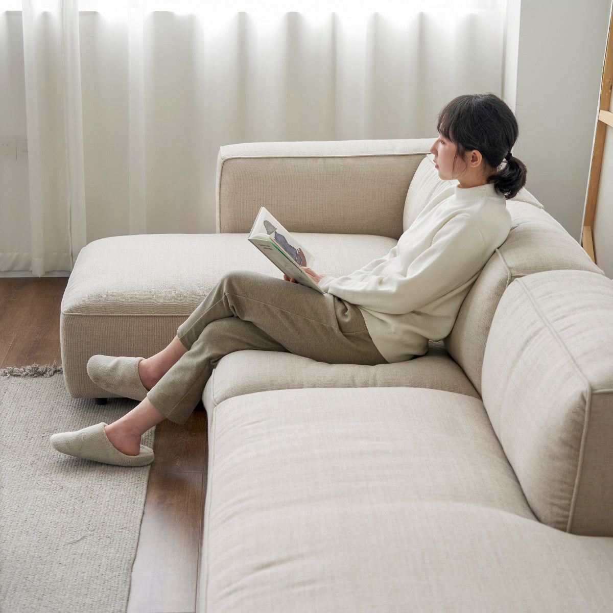 Woman sitting on a beige sectional sofa reading a magazine in a bright living room.
