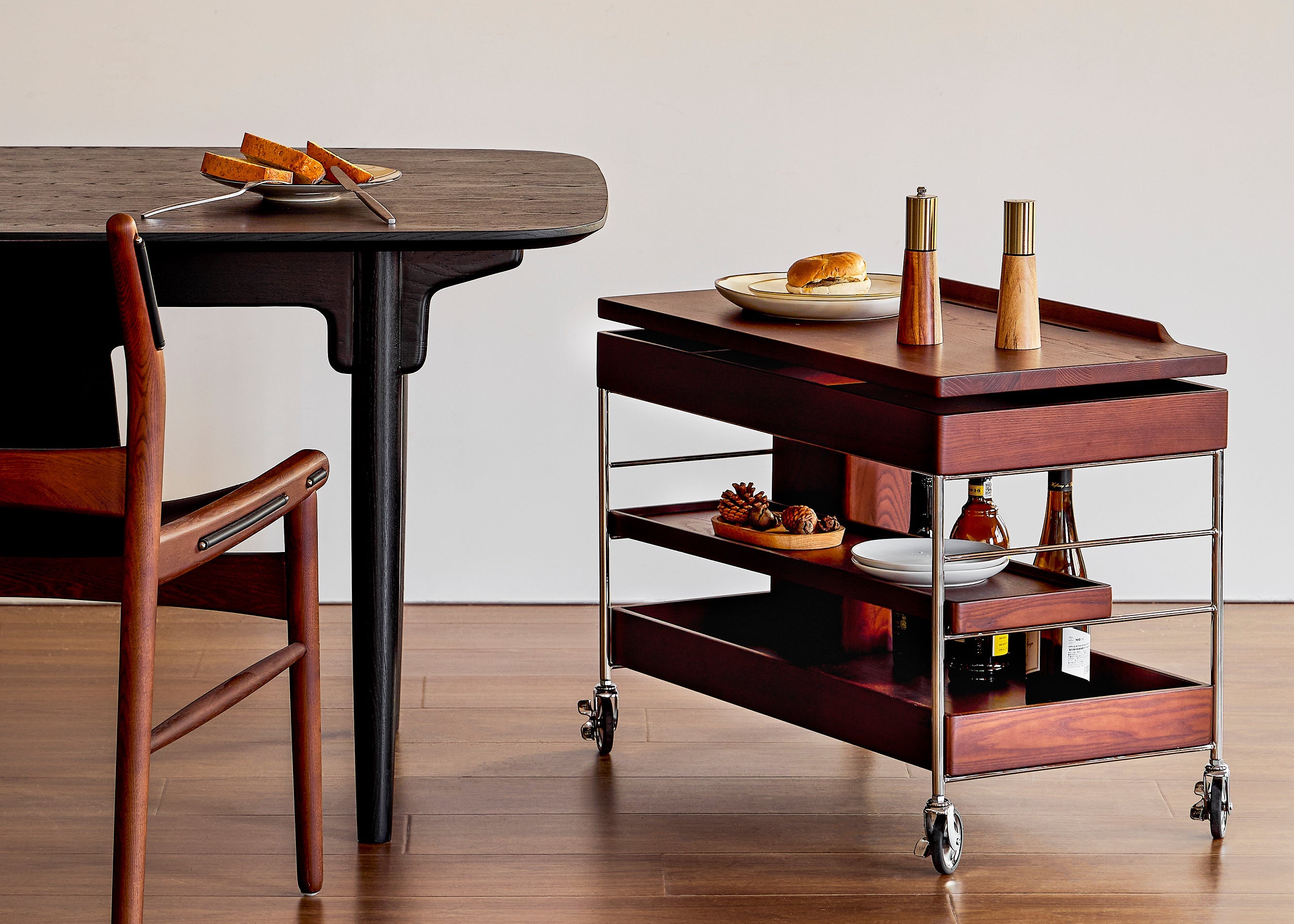 Wooden dining table with chair and rolling cart against a plain wall with a framed picture.