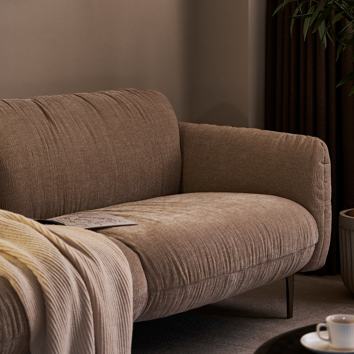 A modern beige upholstered loveseat in a styled living room corner, with a cream knit throw blanket draped over the left side, a plant at right, and a small table with a cup in the foreground.