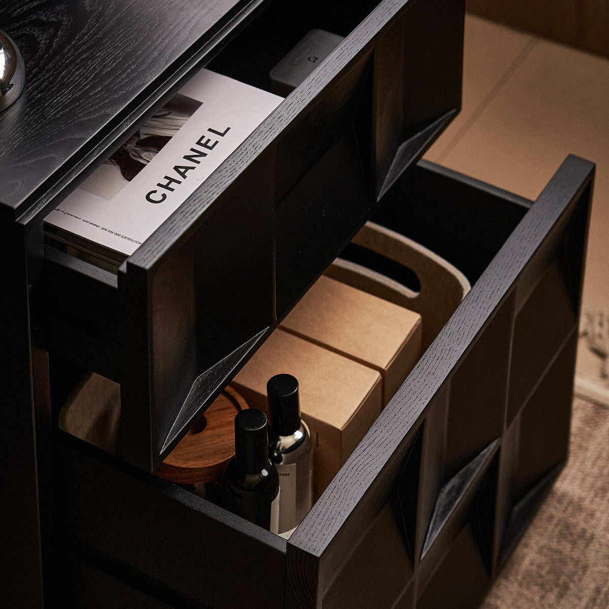 Open drawer of a black nightstand with Chanel magazine and cosmetic items.