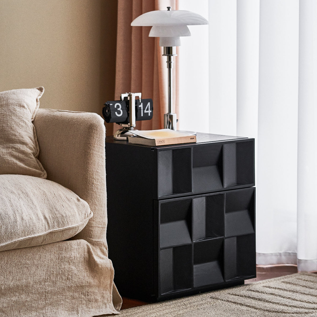 Black geometric nightstand with lamp and books next to a beige sofa in a room with curtains.