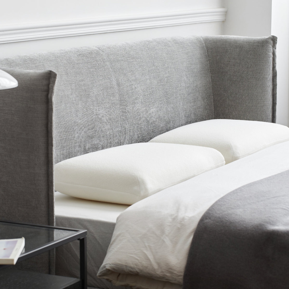 A modern bed with a tall upholstered headboard and footboard in textured gray fabric, styled with white pillows and white bedding. A matching gray throw blanket is draped over the foot of the bed, and a black glass nightstand with a white mushroom-shaped table lamp sits beside it against a white paneled wall.