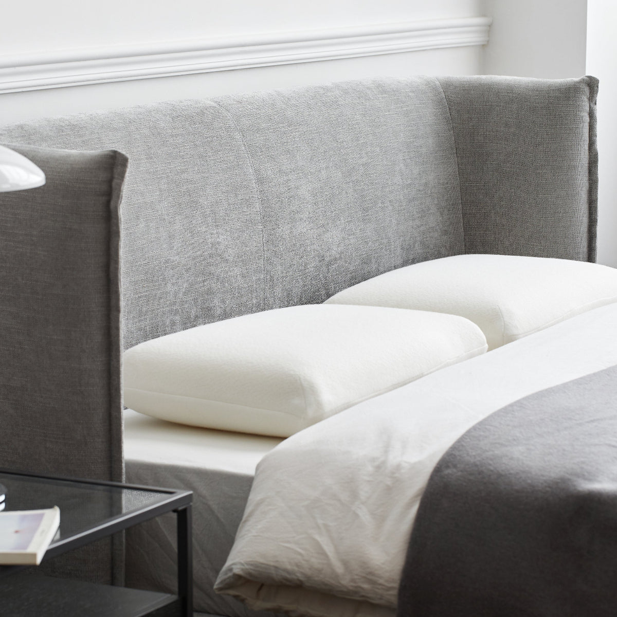 Gray upholstered headboard with white pillows and a glass nightstand with a lamp.