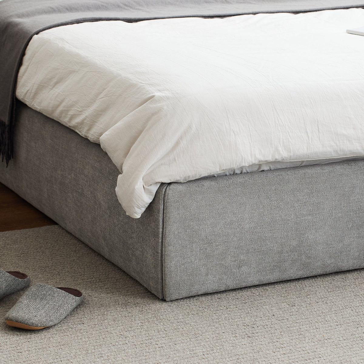 Gray upholstered bed with white bedding and gray slippers on a carpeted floor.