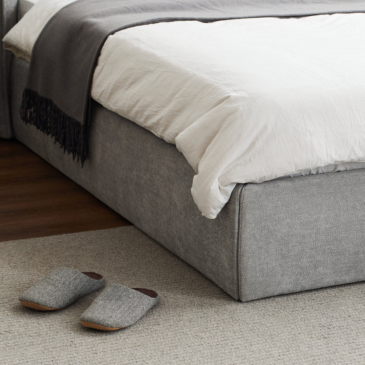 A low-profile upholstered bed frame in light gray fabric, dressed with white bedding and a dark gray throw blanket, with a pair of matching gray slippers on a light rug nearby.