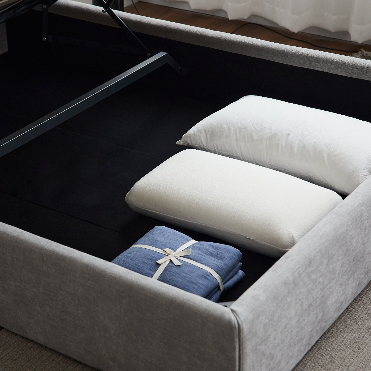 A gray upholstered bed frame with the lift-up mattress platform open, showing a black interior. Two white pillows and a folded blue blanket are placed inside the storage compartment.