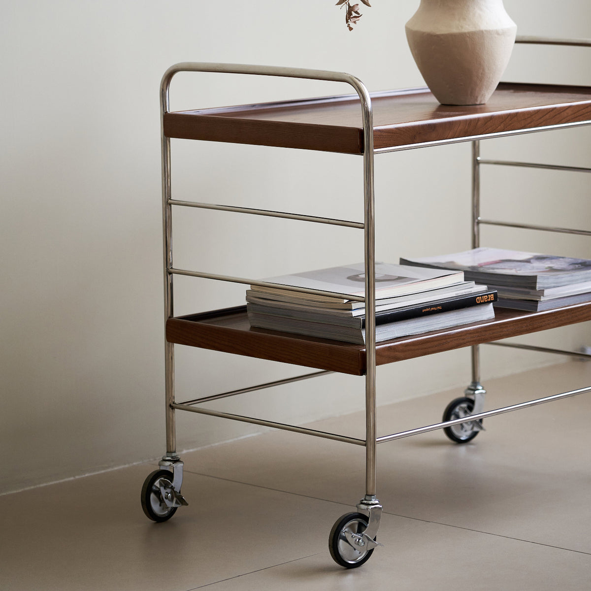 Wooden serving cart with metal frame and wheels, holding a vase and books against a plain wall.