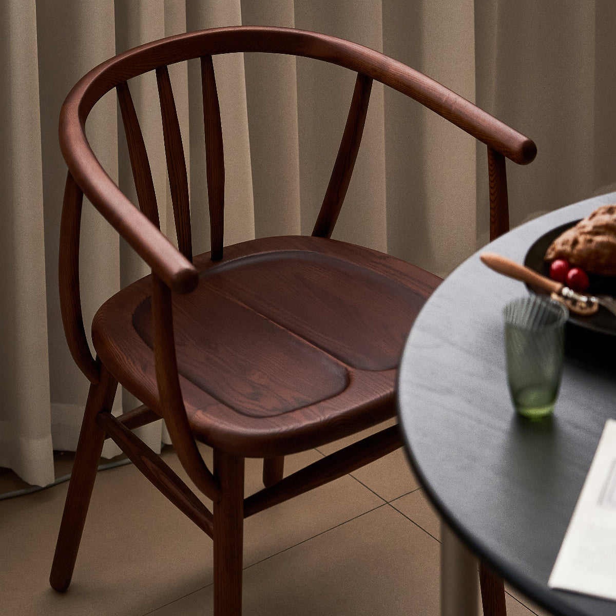 Wooden chair next to a round table with a glass and a plate of food in a softly lit room.