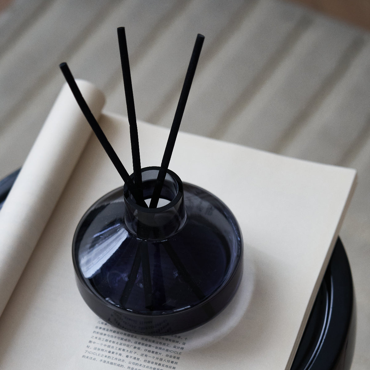 A dark navy blue glass reed diffuser bottle with three black reeds, resting on an open book.