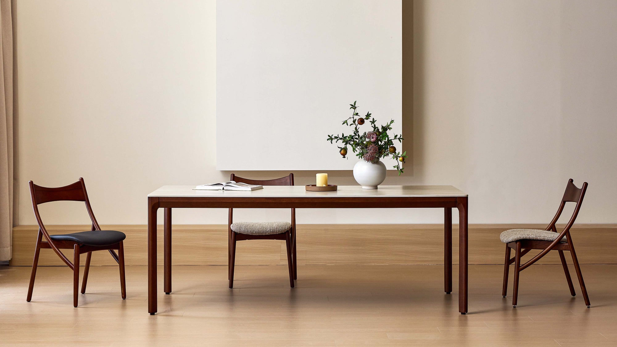A minimalist dining room scene featuring a rectangular wooden dining table with a light top, accompanied by three matching wooden chairs with dark, light, and gray upholstered seats, styled with a white vase of flowers on a light wood floor against a beige wall.
