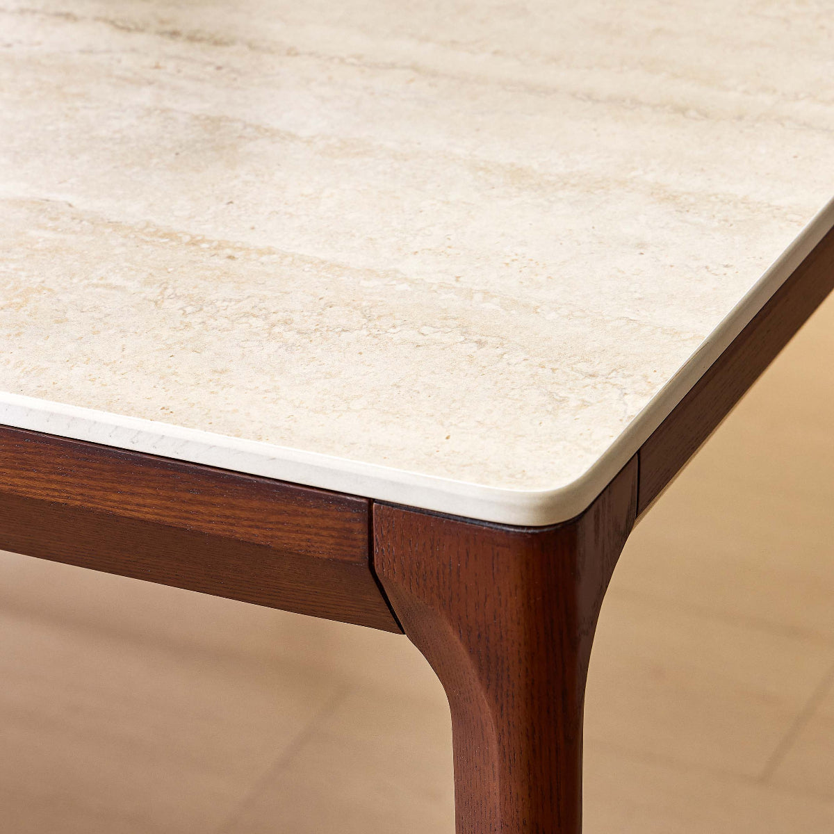 Close-up of a dining table corner showing a light beige stone or marble top with a warm brown wooden frame and leg, set against a light wood floor.