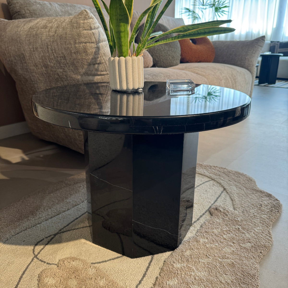 Modern black vase on a table with a plant and decorative pillows in a living room setting.