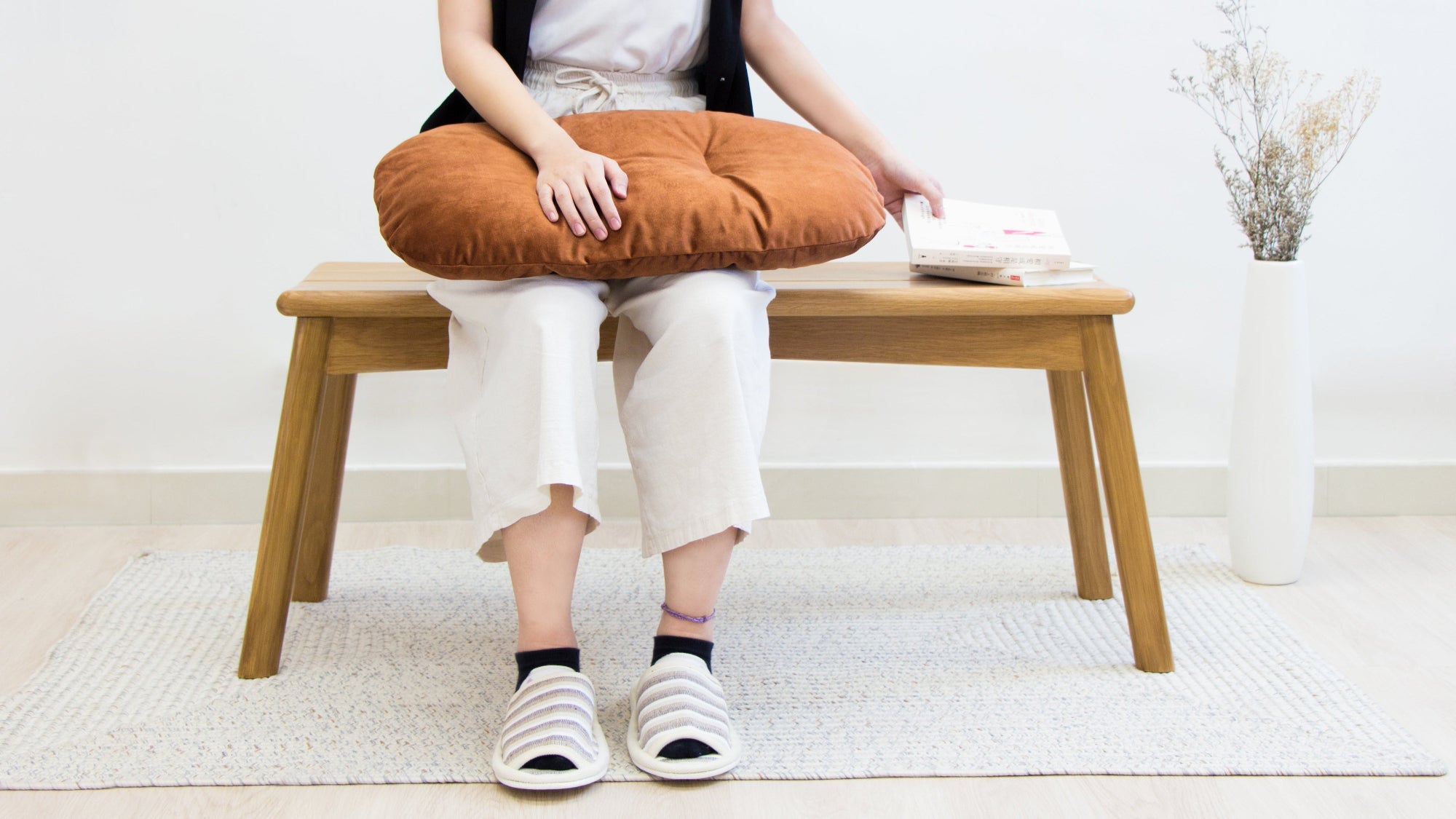 Person sitting on a wooden stool with a brown cushion, wearing white pants and black shoes.