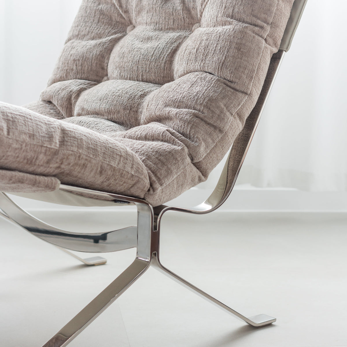 A close-up of a modern lounge chair with a tufted beige fabric cushion and a curved chrome metal frame and base, set against a bright, minimalist white background.