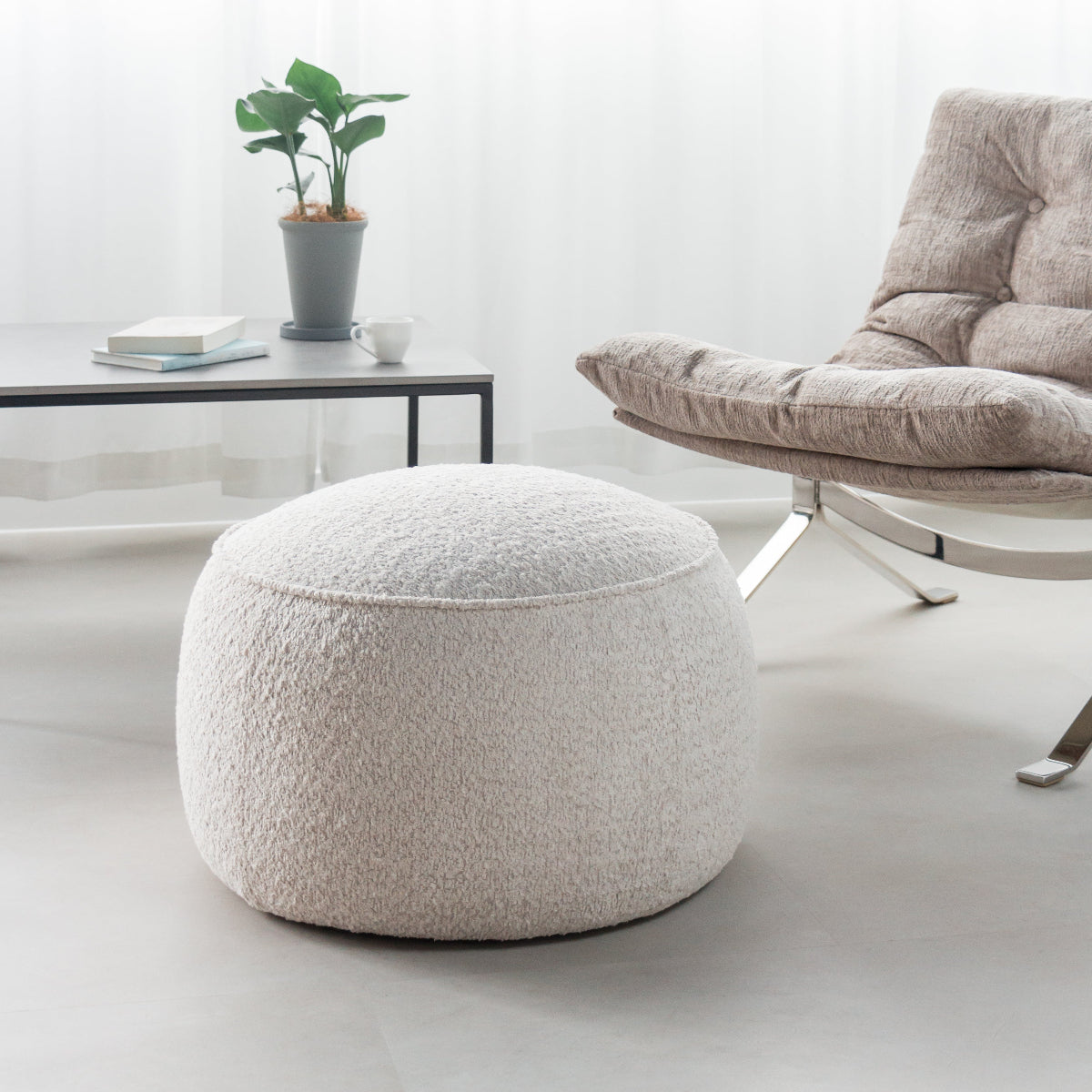 A round, low-profile pouf ottoman upholstered in a textured bouclé fabric in a warm off-white or cream color, styled in a bright, modern living room next to a tufted armchair.