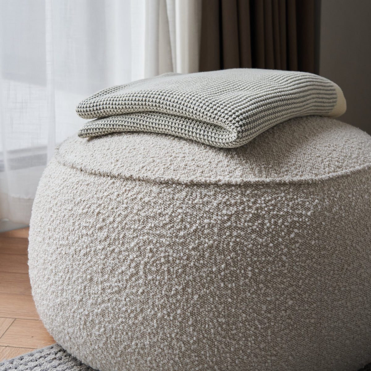 A round, textured pouf ottoman in a light beige bouclé fabric, with a folded knit throw blanket in a gray and white striped pattern resting on top, set in a softly lit room with curtains and wood flooring.