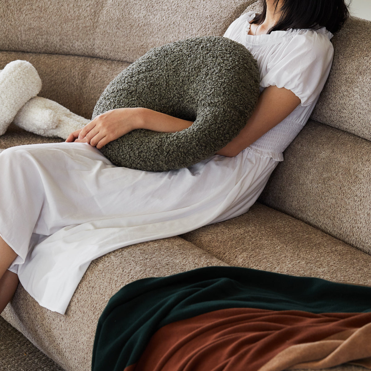 Person sitting on a beige couch with a green textured pillow, wearing a white outfit.