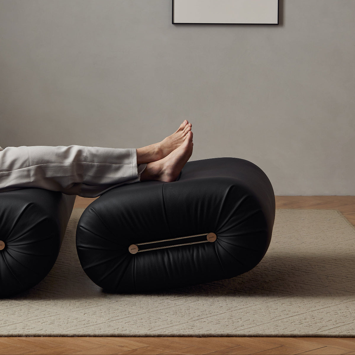 A person's bare feet resting on a black cylindrical floor cushion in a minimalist room with a gray wall and a framed black-and-white print.