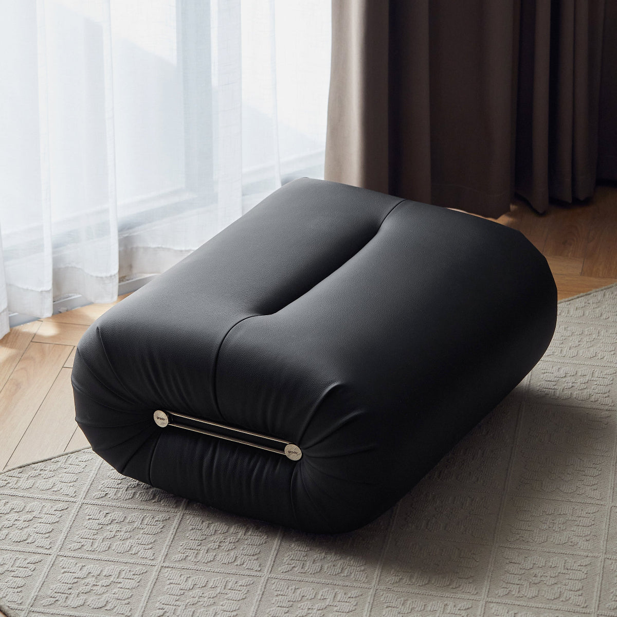 A black tufted floor cushion or ottoman with a metallic bar detail, placed on a textured light rug in a room with sheer white curtains, brown drapes, and a wooden floor.