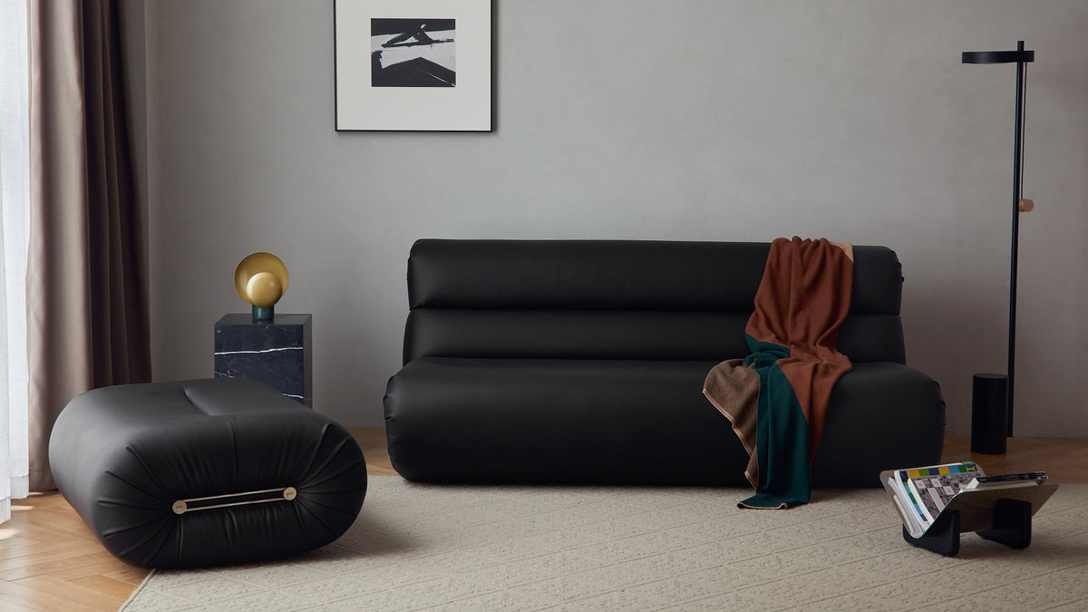A modern black leather sofa in a minimalist living room setting, styled with a rust-colored throw blanket, a black floor lamp, and a black cylindrical ottoman on a beige rug.