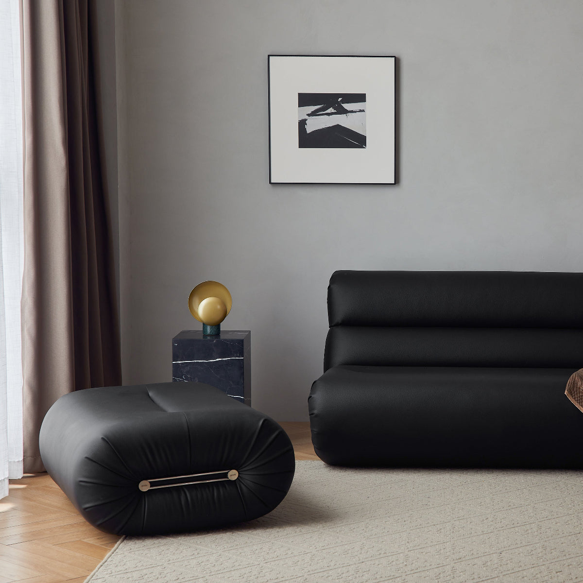 A modern black leather sofa in a minimalist living room setting, styled with a rust-colored throw blanket, a black floor lamp, and a black cylindrical ottoman on a beige rug.