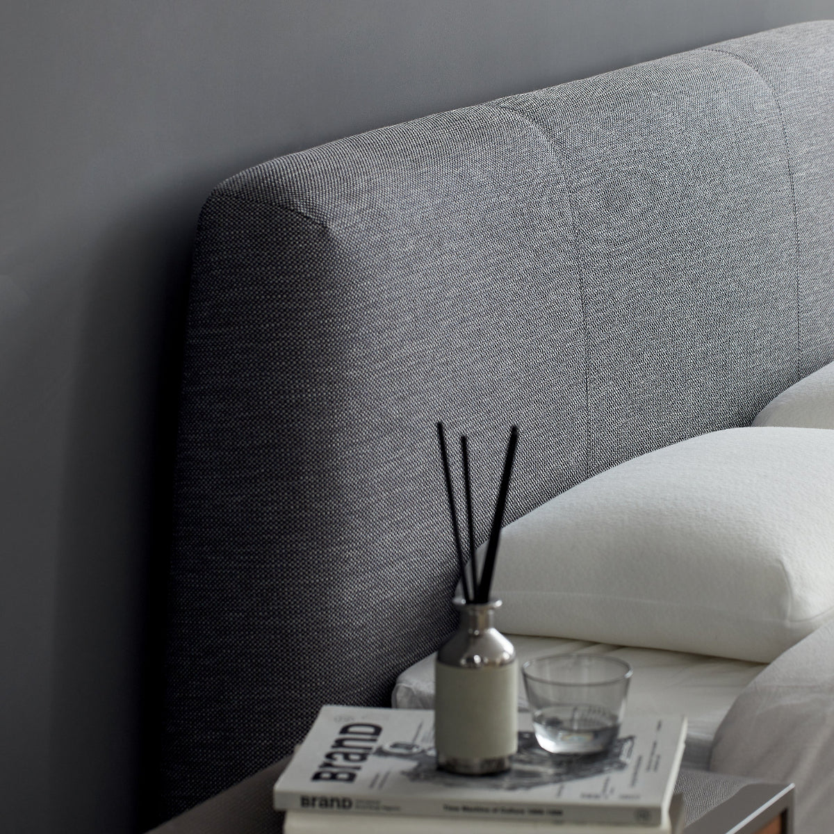 Gray upholstered headboard with a nightstand and decorative items against a gray wall.