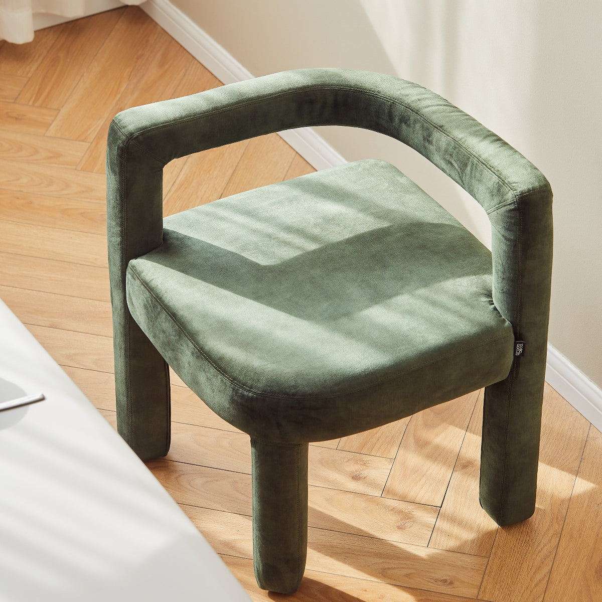 Green velvet chair on a wooden floor with white curtains in the background