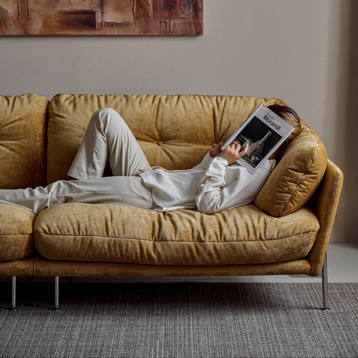 Person lying on a beige sofa reading a magazine in a room with abstract art on the wall.