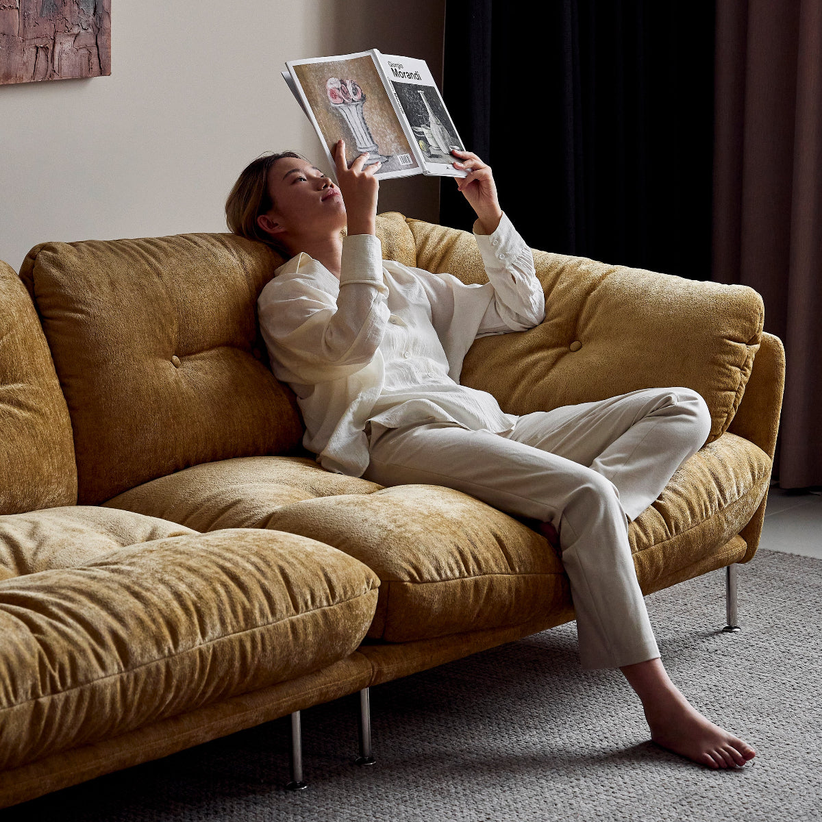 Person sitting on a brown sofa reading a magazine in a room with dark curtains.