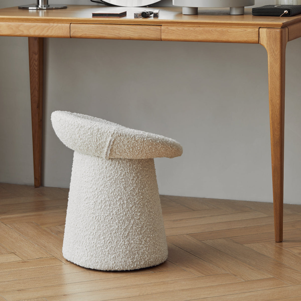 Wooden desk with round mirror, lamp, and decorative items in a room with a textured white stool.