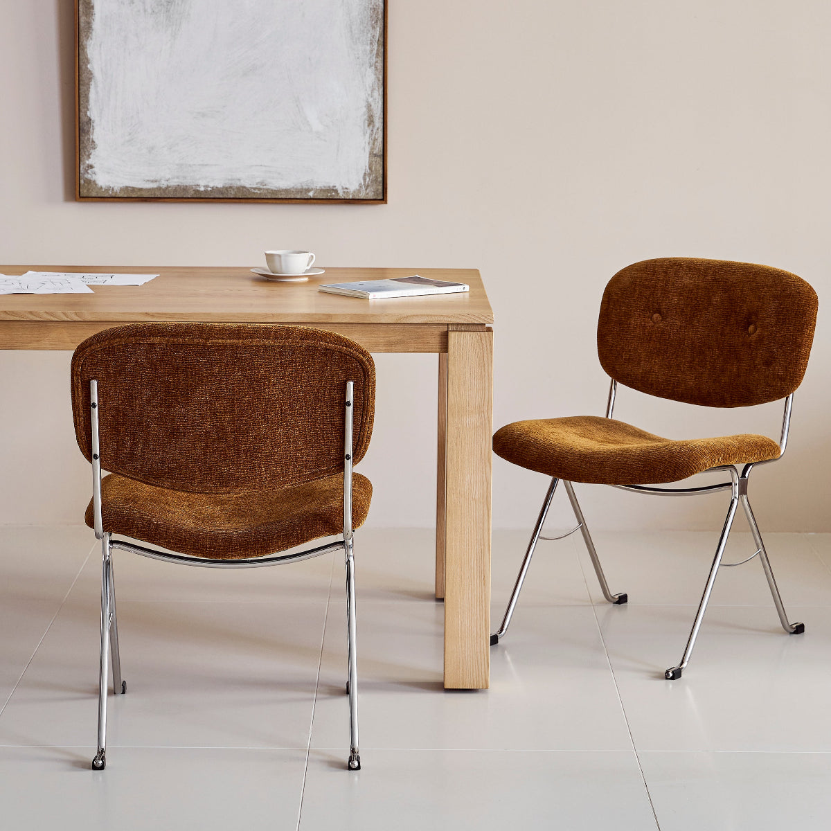 Two brown chairs in front of a wooden table with a white wall and framed abstract art in the background.