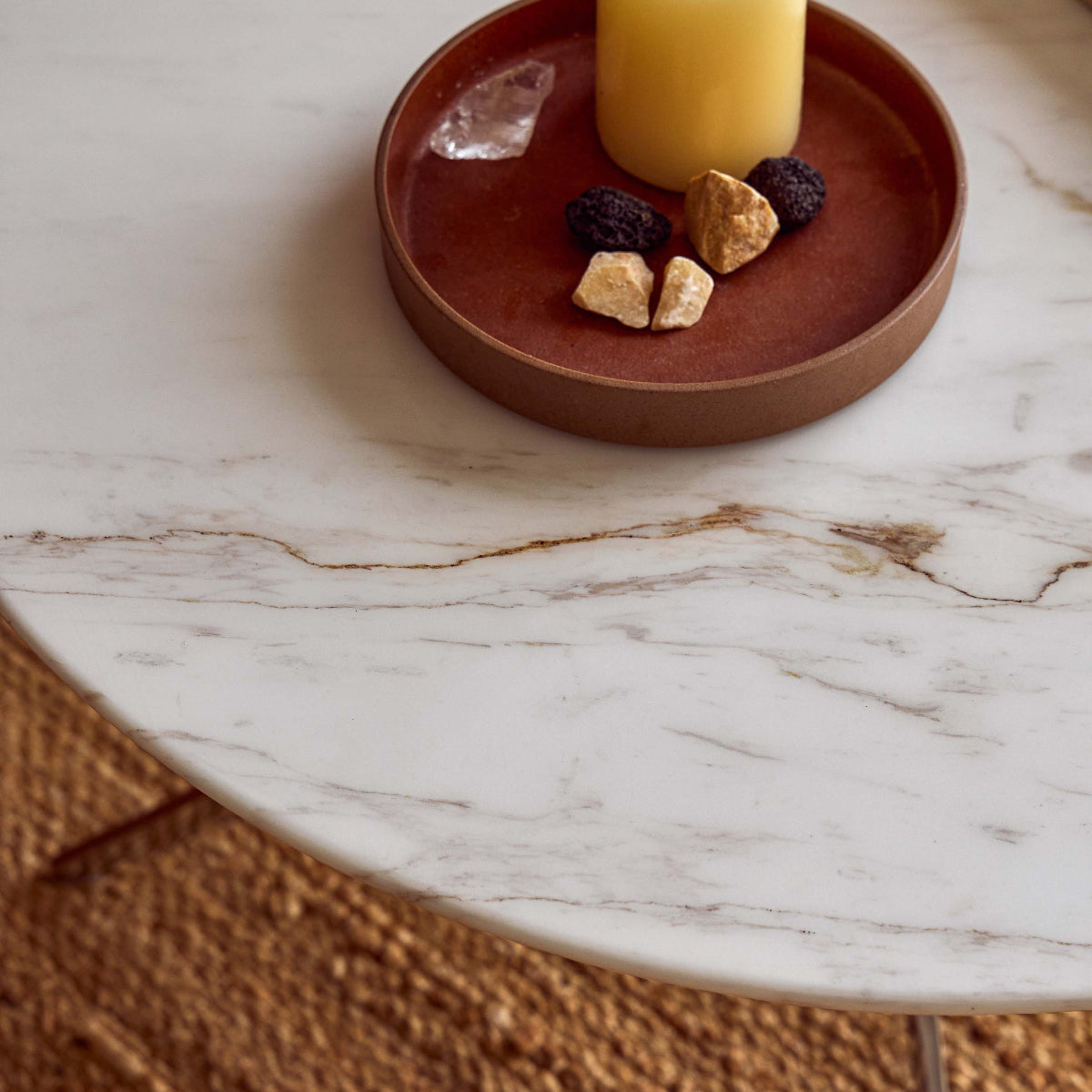 A round brown ceramic tray holding a yellow pillar candle, crystals, and small stones, placed on a white marble table with a woven rug underneath.