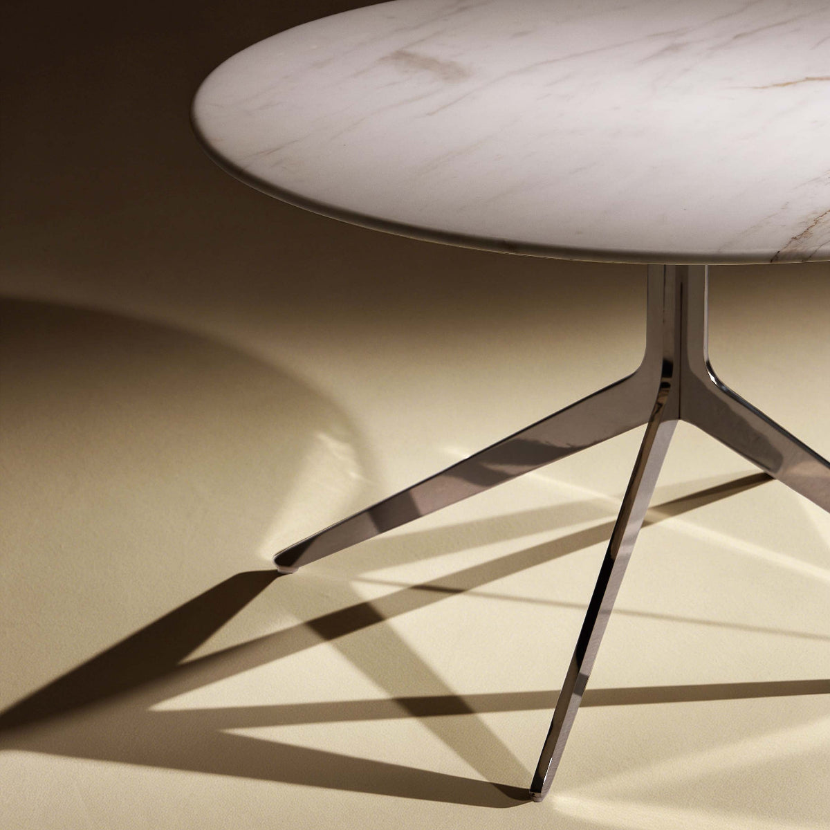 A modern round coffee table with a white marble top and a chrome tripod base, shown in a warm, dramatic studio setting with sharp shadows.