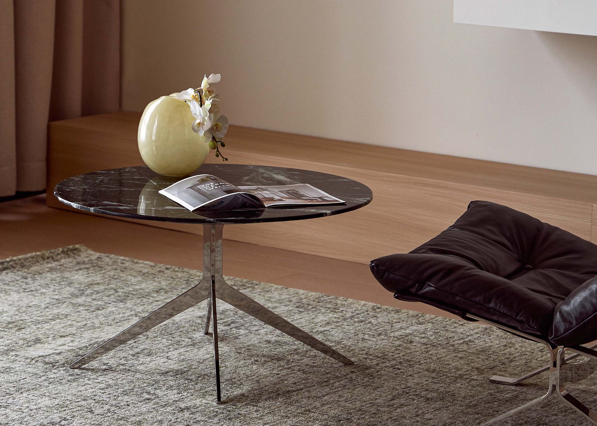 A round glass-top coffee table with a metal tripod base sits on a textured gray rug in a modern living room setting. A pale yellow vase with dried stems and an open magazine rest on the tabletop.