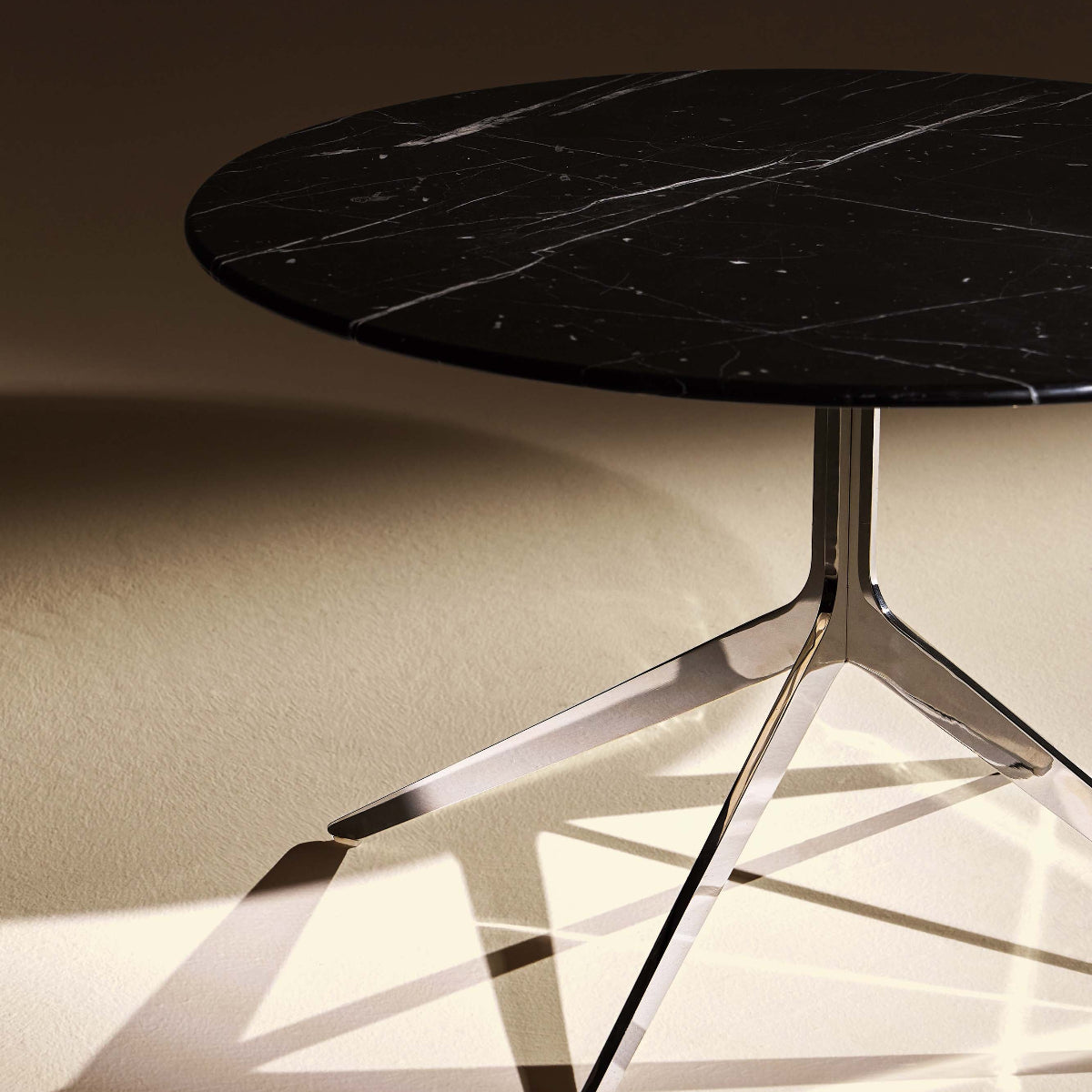 A modern round side table with a black marble top and a polished chrome tripod base, shown in a minimalist room setting with dramatic warm lighting and sharp shadows.