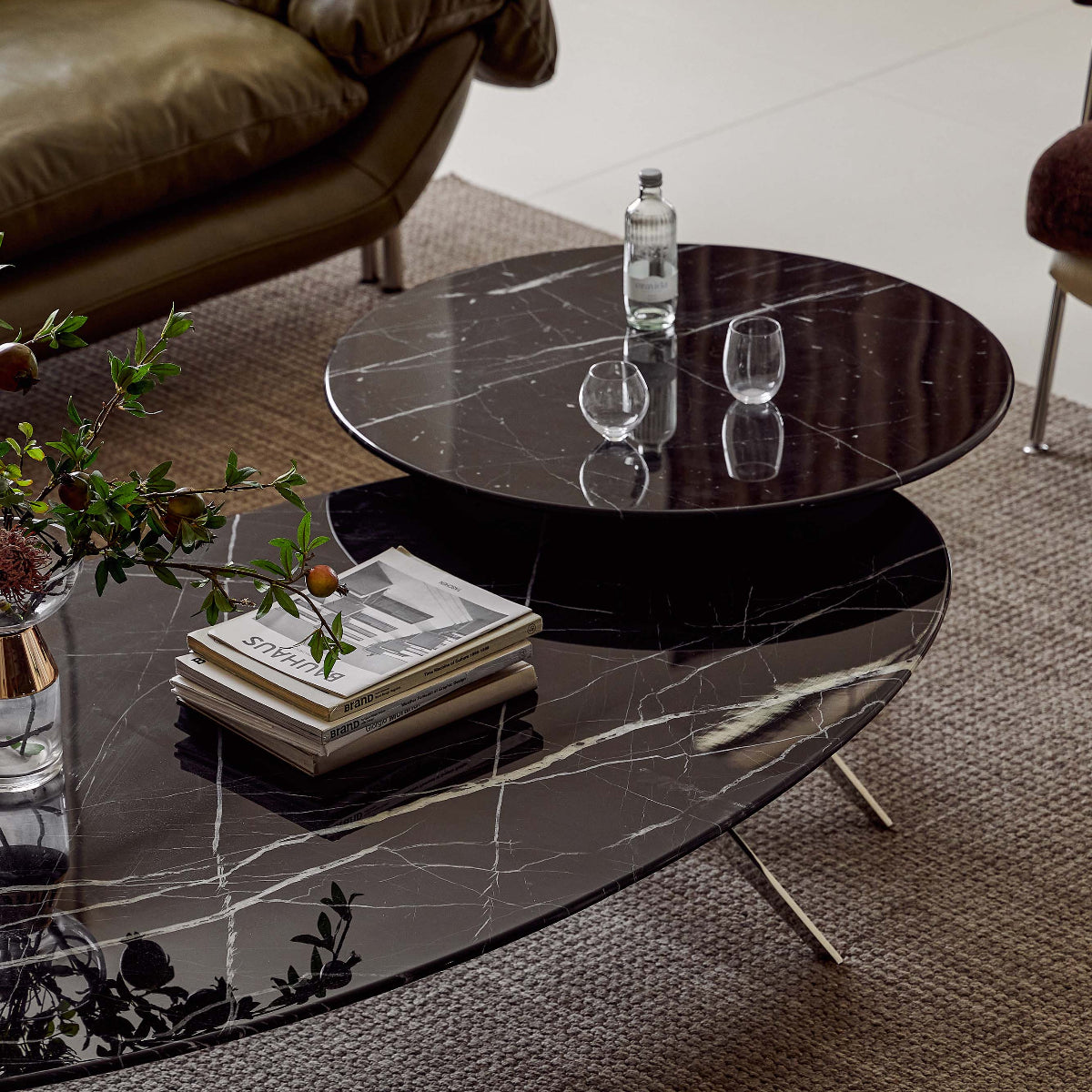 A modern oval coffee table with a black marble surface and thin metal legs, styled in a living room setting with books, a bottle, and glasses on top.