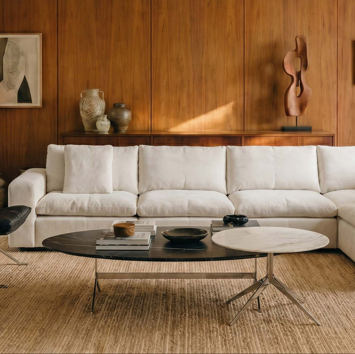 A modern living room featuring a white sectional sofa, two black leather armchairs, a round black coffee table, and a woven beige rug, set against a warm wood-paneled wall.