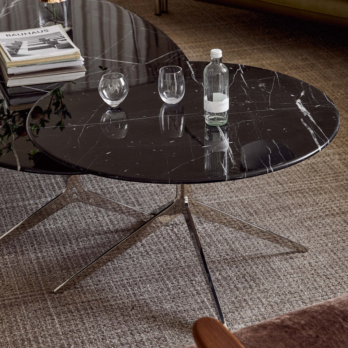 A modern round black marble coffee table with a chrome star base in a living room setting, styled with books, a glass vase, and a water bottle.