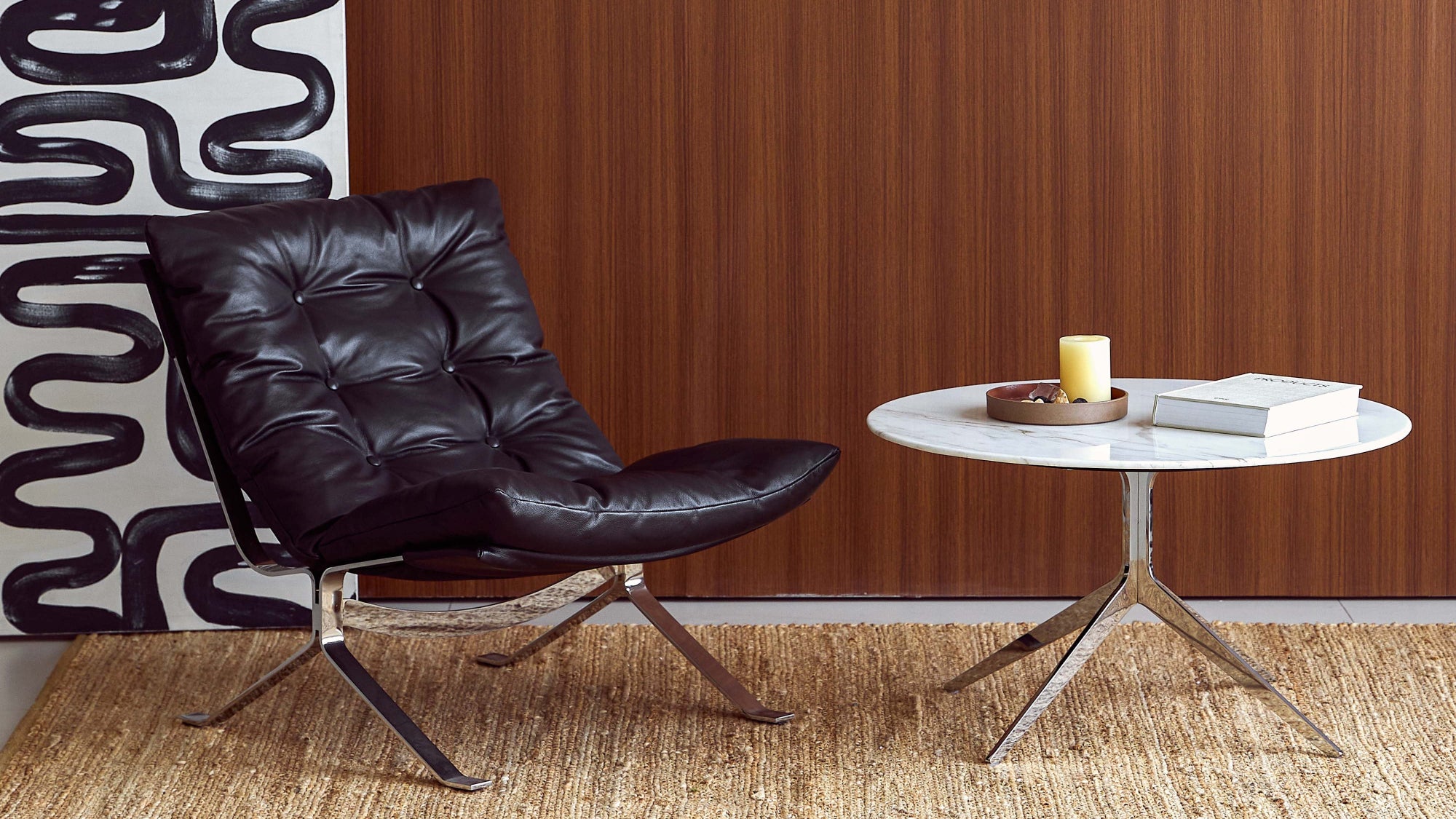 A modern black leather tufted lounge chair sits beside a small round white-top table with metal legs in a styled interior. A large black-and-white abstract wall art piece is visible on the left against a brown wood-paneled wall.