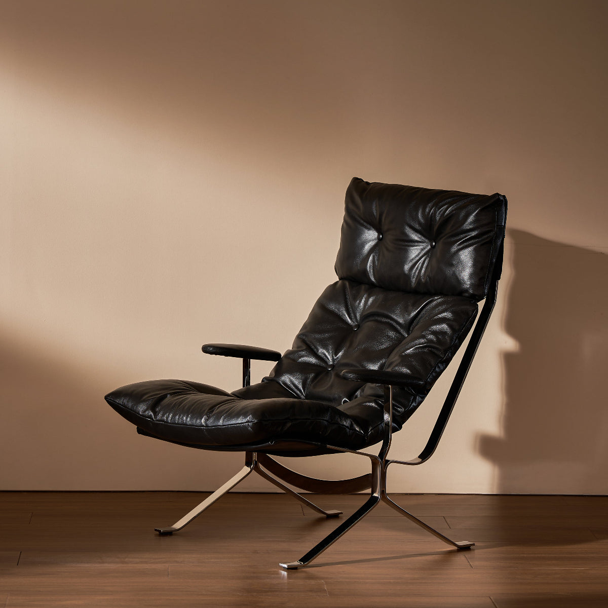 A modern black leather recliner chair with a tufted backrest and padded footrest, supported by a chrome metal tripod base, set against a warm beige wall and dark wood floor.
