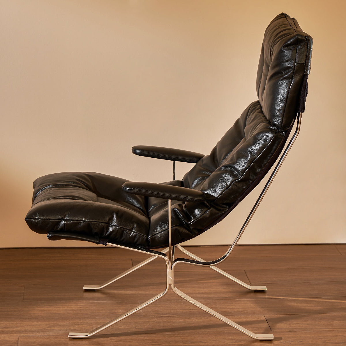 A modern black leather lounge chair with a high back, padded armrests, and a chrome metal star base, set against a warm beige wall and dark wood floor.