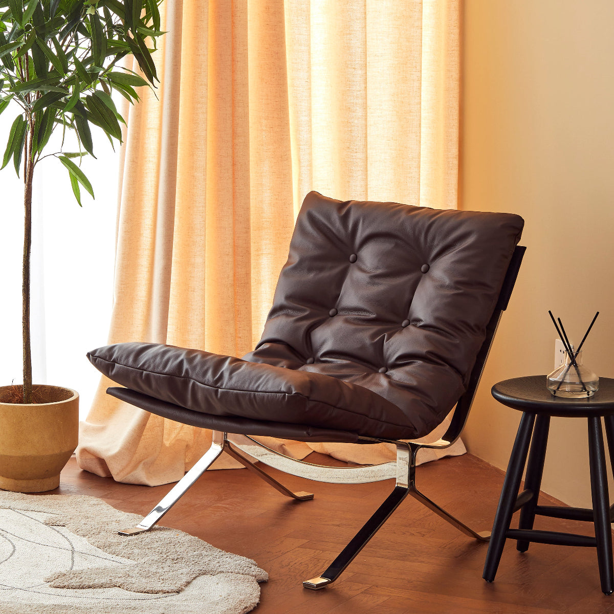 A modern brown leather lounge chair with a tufted cushion and chrome legs, styled in a warm room with beige curtains, a potted plant, and a small side table.