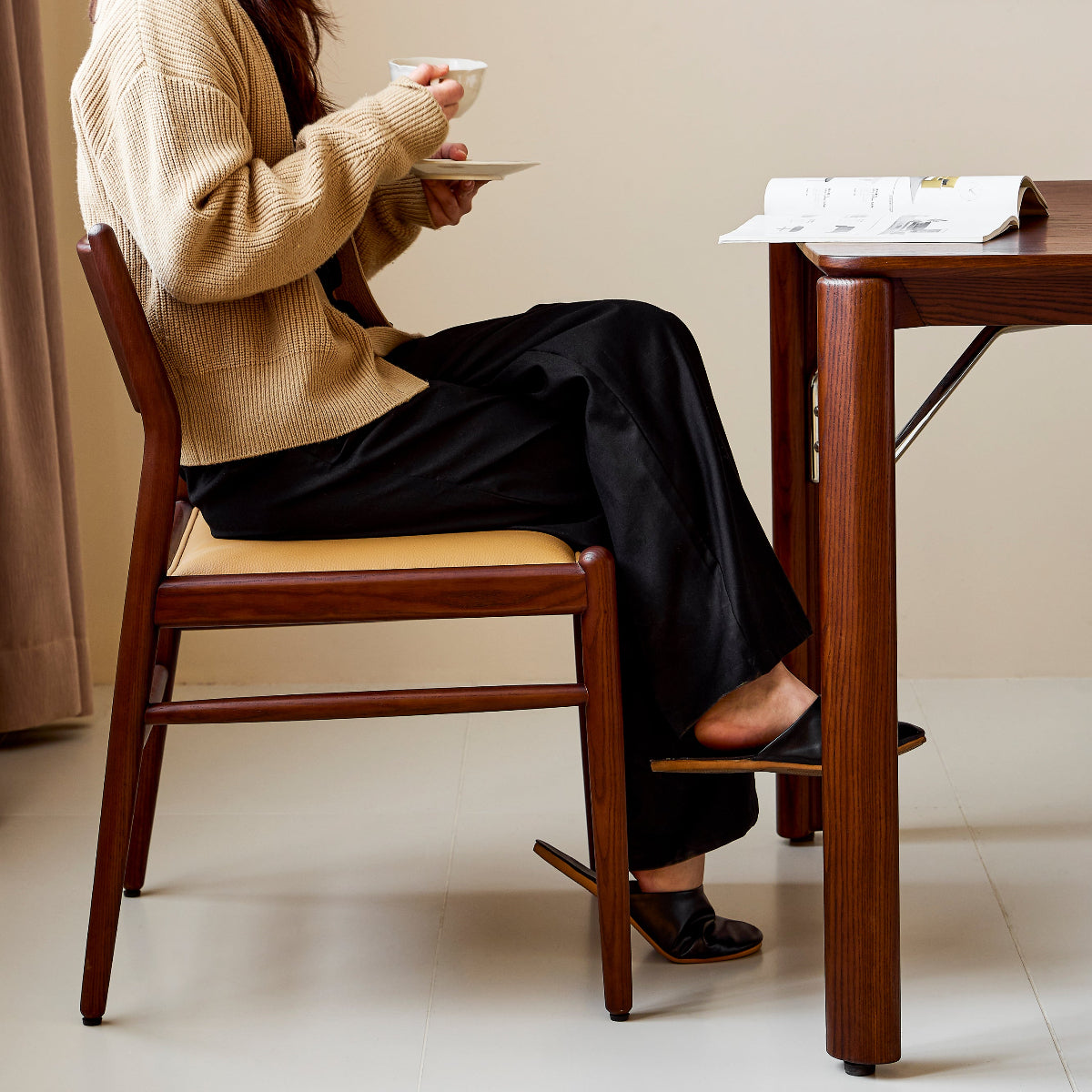 Woman sitting at a wooden table holding a cup, with a painting on the wall in the background.