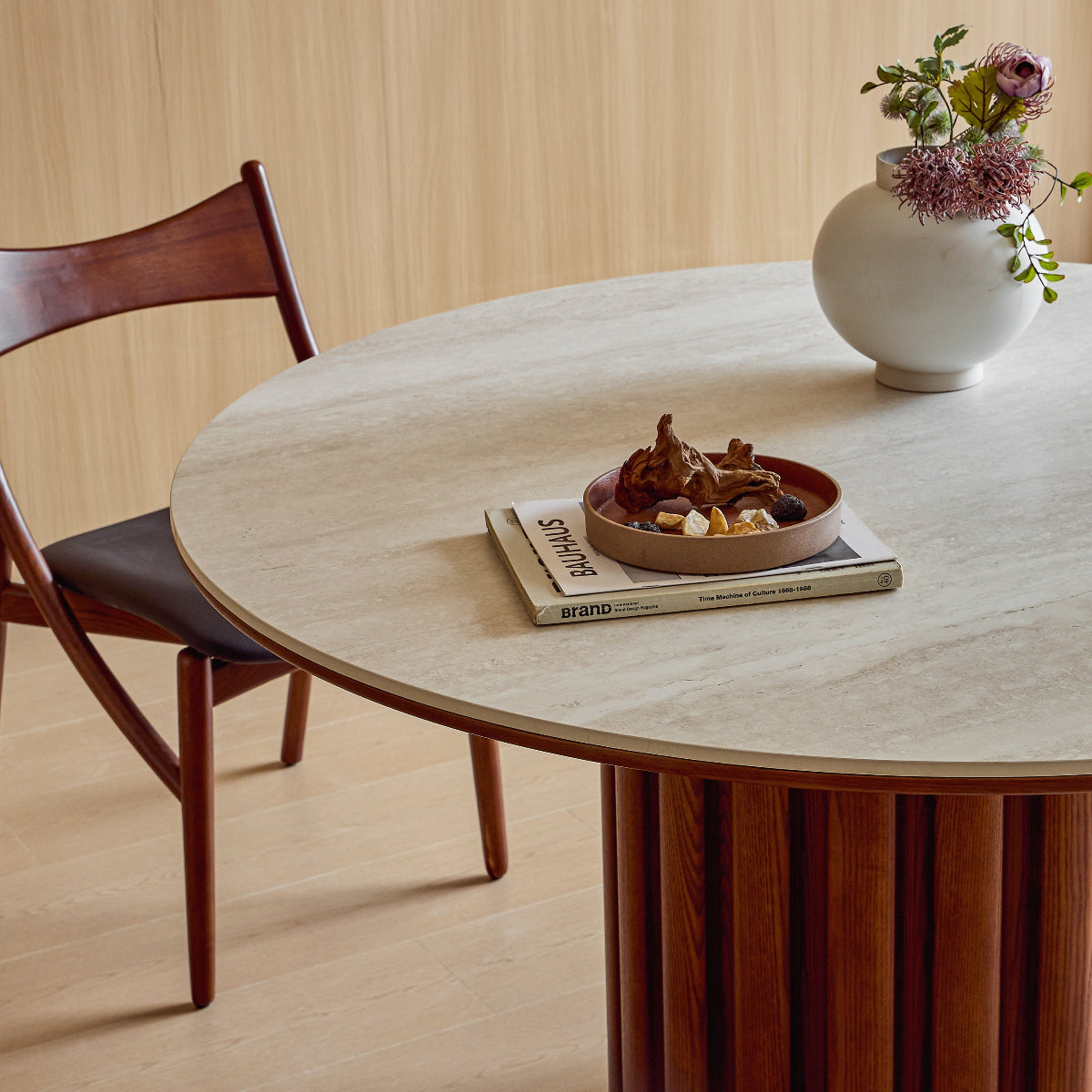Round wooden dining table with a vase of flowers and a bowl on top, in a room with wooden flooring and a chair.