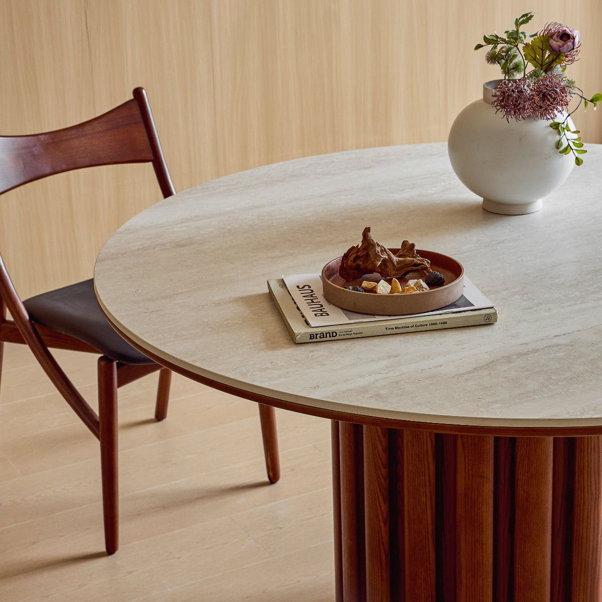 Round wooden dining table with a vase of flowers and a bowl on top, in a room with wooden flooring and a chair.